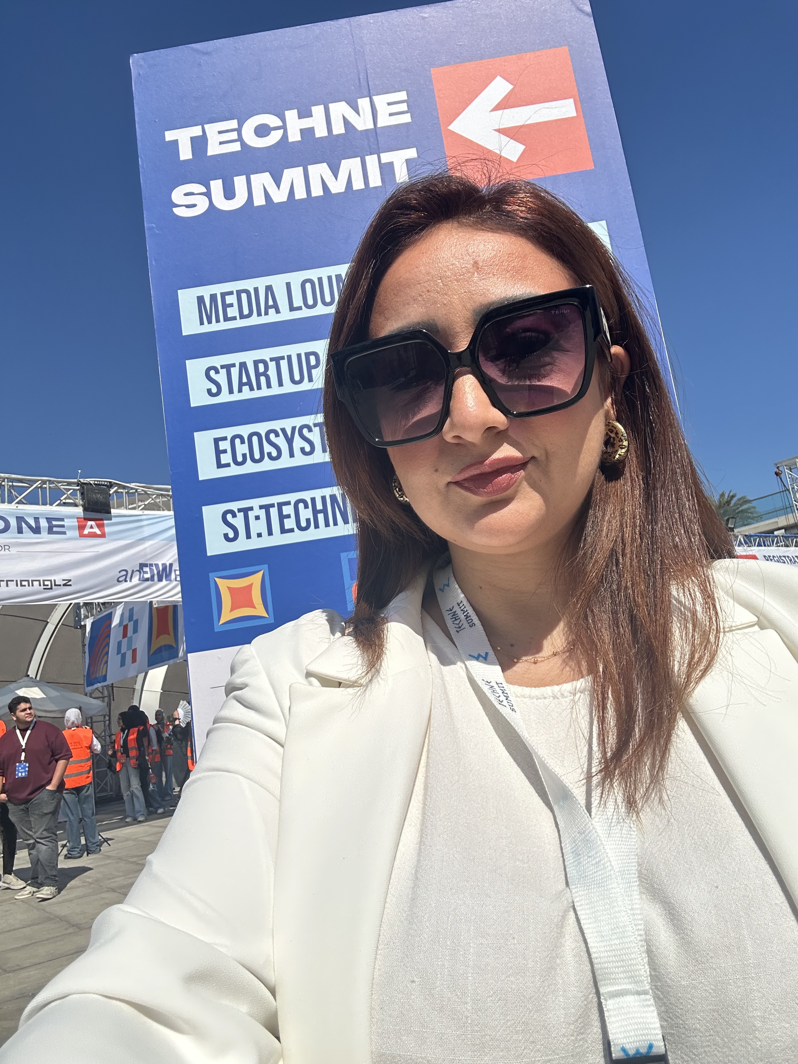 Mai in a white blazer and sunglasses beside the outdoor Techne Summit signage in 2025