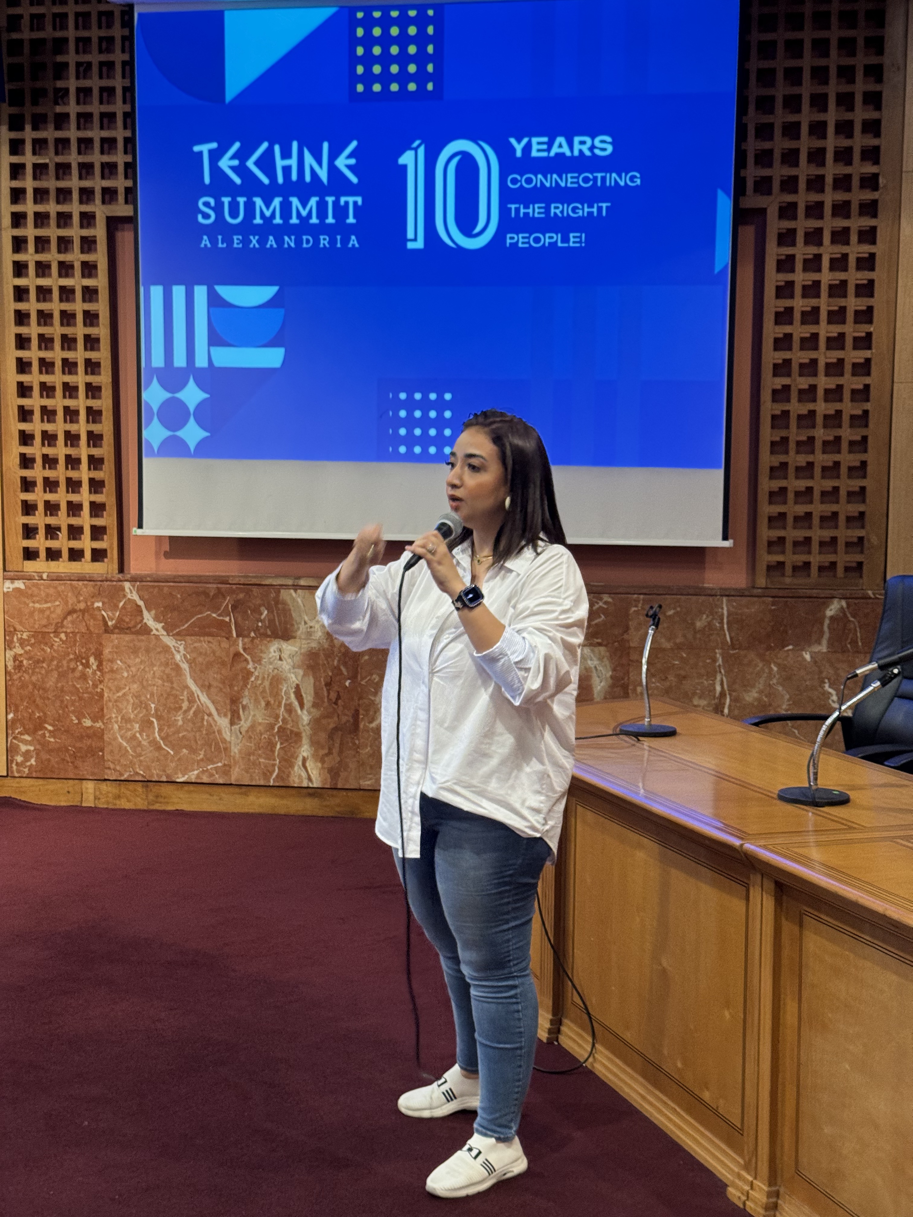 Mai speaking in a white shirt in front of the blue Techne Summit Alexandria screen
